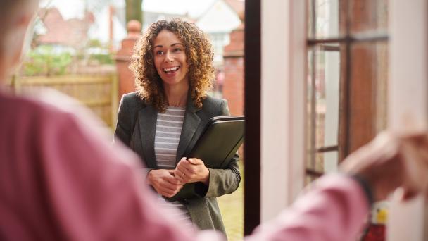 Unidentifiable person opening their front door in the foreground, and a woman with folder working as a survey interviewer smiling.